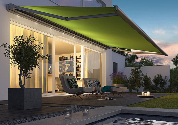 Die Terrassenmarkise Cassitta 2 in hellgrün in der Abenddämmerung mit schönen LEDs. Sicht aus dem Garten auf Wohnhaus. Klicken um zur Cassitta 2 Info zu gelangen.